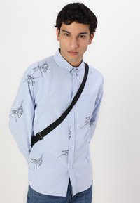 Light blue shirt with embroidered black crickets, button-down collar, patch pocket, and long sleeves. Worn with a black crossbody strap.