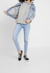 Sudadera con capucha gris debajo de una chaqueta de mezclilla azul claro, combinada con pantalones vaqueros ajustados azul claro y zapatillas negras con detalles blancos.