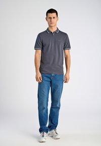 Dunkelgraues Poloshirt mit Reißverschluss am Kragen, weißen Akzenten an den Bündchen, kombiniert mit blauen Jeans und weißen Sportschuhen.