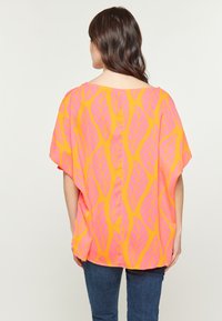 Maglietta arancione brillante con un motivo a foglia in rosa. Vestibilità ampia con maniche corte e scollo ampio. Abbinata a jeans blu scuro.
