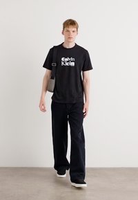 Juodas marškinėliai su balta "Calvin Klein" logo, derinami su laisvomis juodomis kelnėmis ir juodais sportbačiais, laikant mažą pilką petinę rankinę.