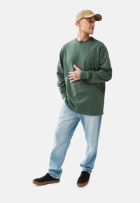 Långärmad grön skjorta med rund hals och logotyp, kombinerad med ljusblå jeans och svarta sneakers. En avslappnad keps lägger till en enkel detalj som accessoar.