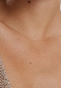 Primer plano de la piel que muestra una textura suave en un tono bronceado claro, con varios lunares pequeños visibles en la zona del cuello.