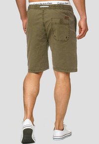 Olive green shorts with elastic waistband, back pocket detail, and stitching accents, paired with white sneakers. Suitable for casual wear.