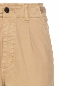 Gros plan de la ceinture d'un pantalon beige avec un bouton noir, des passants de ceinture, une poche latérale et des détails de couture visibles.