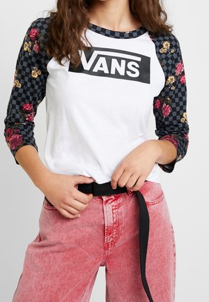 Osoba nosząca białą koszulkę Vans z czarnymi kratką i kwiatowymi rękawami, różowe wyprane jeansy, poprawiająca czarny pasek.
