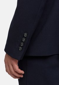 Granatowy rękaw marynarki wykonany z teksturowanej tkaniny, z czterema czarnymi guzikami z motywami kwiatowymi oraz kontrastującym przeszyciem przy mankiecie.