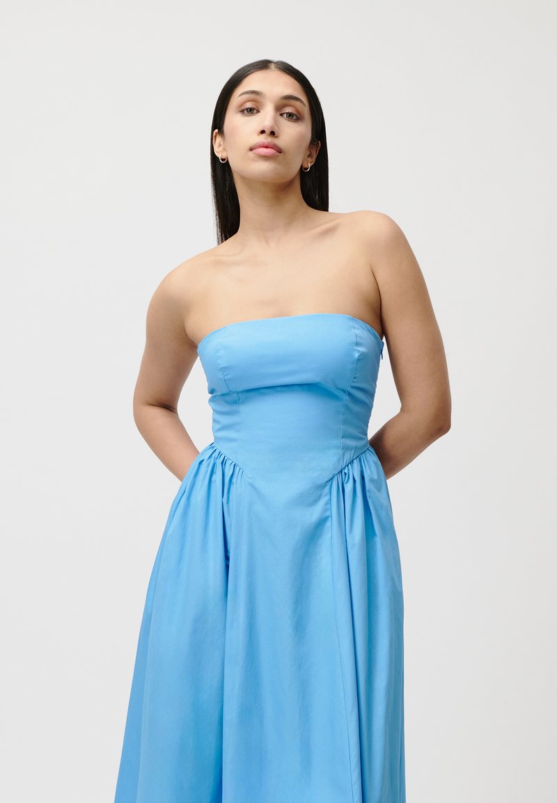 Bezramieniowa niebieska sukienka charakteryzuje się dopasowaną górą, marszczonym dołem i gładkim, miękkim materiałem. Brak wzorów i widocznych detali metalowych.