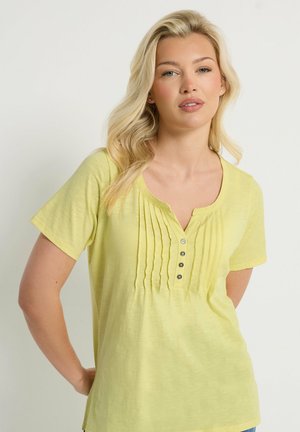 Femme blonde aux cheveux ondulés portant un haut à manches courtes jaune pâle avec des plis plats à l'avant et quatre boutons, debout devant un fond uni.