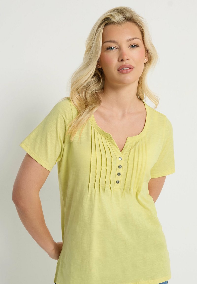 Blondynka o falowanych włosach, ubrana w bladożółty top z krótkim rękawem z zakładkami z przodu i czterema guzikami, stojąca na jednolitym tle.