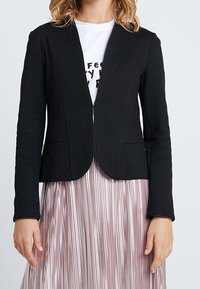 Czarna marynarka o dopasowanym kroju, z otwartym przodem i dwoma przednimi kieszeniami, zestawiona z plisowaną różową spódnicą. Materiał wydaje się gładki i lekko elastyczny.