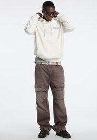 Roomkleurige hoodie met logo, gedragen over een grijs shirt. Bruine cargobroek met zijzakken en een losse pasvorm. Zwarte zonnebril en donkere schoenen.