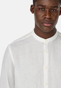 Camicia bianca in lino con colletto alla cinese, caratterizzata da un tessuto testurizzato e apertura con bottoni. Design semplice e materiale leggero e traspirante.
