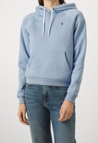 Sudadera azul claro con un bolsillo canguro, presenta una capucha con cordones, puños de canalé y un pequeño logo en el pecho. Tejido suave y ajuste casual.