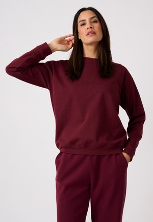 Donna con capelli scuri che indossa una felpa bordeaux e pantaloni coordinati, in piedi con una mano dietro la testa e l'altra in tasca.