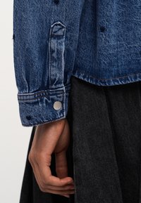 Χέρι που ακουμπά στο πλάι, φορόντας τζιν μπουφάν με μανίκι με κουμπί πάνω από σκούρα φούστα ή παντελόνι.