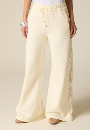 Žena nosící krémové široké kalhoty s šňůrkovým pasem a flitrovými bočními pruhy, doplněné béžovými botami a stříbrným náramkem.