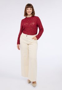 Rotes Spitzenoberteil mit langen Ärmeln und floralem Muster, kombiniert mit weiten cremefarbenen Hosen. Das Model steht mit den Händen in den Taschen und trägt hellfarbene High Heels.
