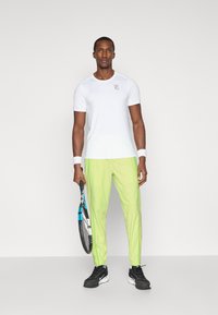 Camiseta atlética blanca de manga corta con logo, combinada con pantalones ajustados verdes brillantes. Zapatillas deportivas negras. Sosteniendo una raqueta de tenis.