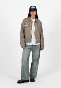 Kvinde iført en taupejakke, hvid stribet skjorte, løse lyseblå jeans, mørke sko og en marineblå baseballkasket, stående foran en hvid baggrund.