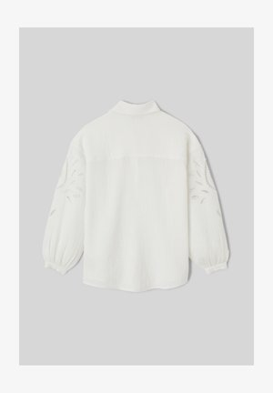 Blouse blanche à manches longues avec manches bouffantes et motifs floraux découpés sur les bras, présentée de dos sur un fond gris.