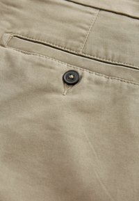 Beige szövet, sima textúrával, fekete gomb részlettel és kis zsebbel. Az éleken látható varrt varrások.