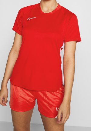 Person, die ein rotes Nike T-Shirt mit kurzen Ärmeln und passende rote Shorts mit weißen Seitenteilen trägt, steht vor einem schlichten Hintergrund.