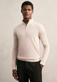 Hellrosa Strickpullover mit einem Viertelreißverschluss-Kragen und langen Ärmeln, kombiniert mit dunklen Hosen. Weiche Textur und figurbetonter Schnitt.