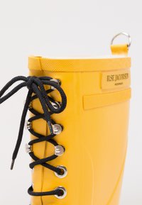 Botte en caoutchouc jaune avec des lacets noirs, des œillets en métal et une étiquette orange portant le nom « ILSE JACOBSEN » en relief. Texture lisse avec un haut arrondi.