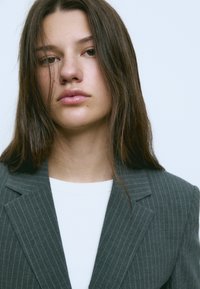 Siva blazer z ozkimi črtami čez belo majico, s klasičnim ovratnikom in gladko teksturo, z ravnimi robovi in minimalnimi detajli.