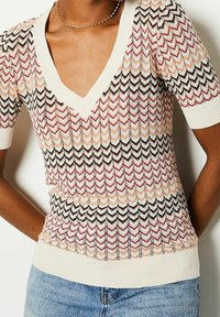 V-neck knit top with short sleeves featuring a chevron pattern in cream, black, red, and peach tones, worn with blue jeans.