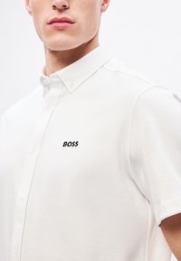 Balti polo marškinėliai su sagomis ant apykaklės, pagaminti iš tekstūruotos medžiagos, kairėje krūtinės pusėje siuvinėtas juodas „BOSS“ logotipas.