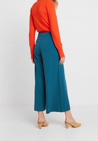 Tealfärgade vida byxor av mjukt tyg, kombinerat med en orange stickad tröja. Modellen har på sig beige klackskor.