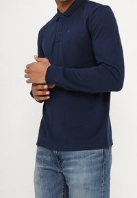 Man i en marinblå långärmad polo med knappar, i kombination med ljusblå jeans, mot en vit bakgrund.