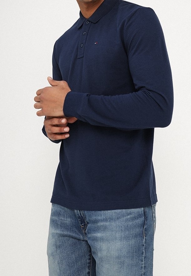 Man i en marinblå långärmad polo med knappar, i kombination med ljusblå jeans, mot en vit bakgrund.