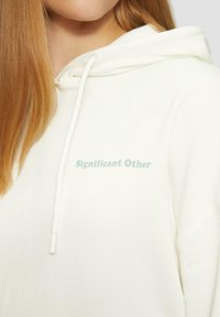 Witte hoodie met een trekkoord, gemaakt van zachte stof, met "Significant Other" in lichtblauw gedrukt. Close-up van het detail aan de voorkant.
