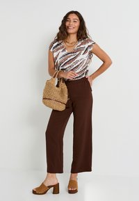Pantalones de pierna ancha marrones, blusa estampada con un patrón abstracto en blanco y marrón, sandalias de plataforma color beige y bolso de mimbre color beige.