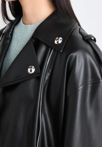 Giacca biker in pelle nera con collo ampio, accessori in argento e cerniera diagonale. Indossata sopra un maglione di maglia azzurro chiaro.