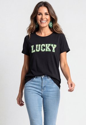 Donna sorridente con capelli mossi indossa orecchini verdi con perline, t-shirt nera con "LUCKY" in motivo verde e jeans azzurri su sfondo semplice.