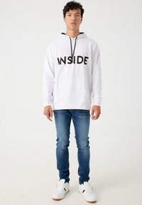 Sudadera blanca con capucha y la palabra "INSIDE" impresa en negro en el pecho, combinada con jeans azules y zapatillas blancas con acentos negros.