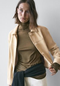 Beige jakke med runde knapper over en mørkegrøn rullekrave og sort strikket sjal, der viser glatte teksturer og tætsiddende design.