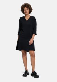 Schwarzes Kleid mit 3/4-Ärmeln, V-Ausschnitt und einem schwingenden Rock. Kombiniert mit schwarzen klobigen Schuhen. Glatte Textur mit einem schlichten Design.