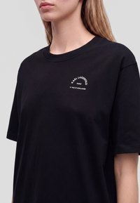 Γυναίκα φοράει μαύρο t-shirt Karl Lagerfeld με λευκό λογότυπο στο αριστερό στήθος, μπροστά σε ουδέτερο φόντο.