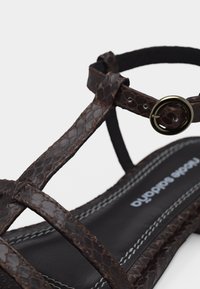 Σανδάλι από σκούρο καφέ δέρμα με υφή, λουρί αστραγάλου με στρογγυλή μεταλλική πόρπη, μαύρο ραμμένο πέλμα και στενό σχέδιο T-λουριού.