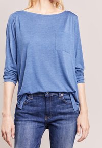 Vrouw in een blauwe lange mouw shirt met een borstzak en blauwe jeans, staande met haar armen ontspannen langs haar zij.