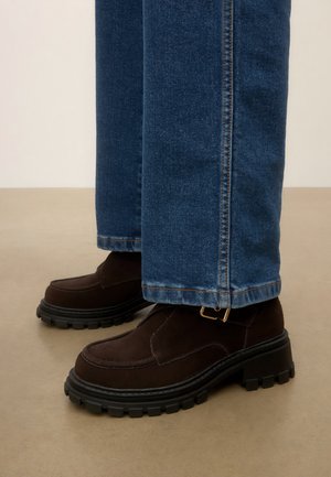 Mocassins en daim brun foncé avec semelles épaisses noires portés avec un jean droit bleu sur un sol beige.