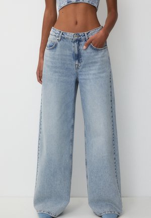 Kobieta ubrana w jasnoniebieskie jeansy z wysokim stanem i szerokimi nogawkami oraz krótki top, z jedną ręką w kieszeni na jednolitym tle.
