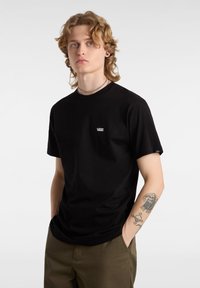 Czarny, krótki rękaw t-shirt wykonany z bawełny, z białym logo "VANS" na klatce piersiowej. Model ma na sobie oliwkowe spodnie, a na ramionach widoczne są tatuaże.