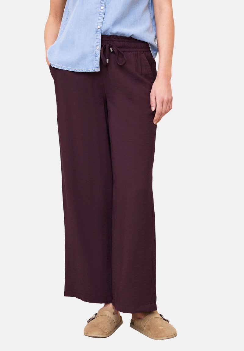 Femme portant un pantalon ample violet foncé à cordon, une chemise sans manches bleu clair boutonnée et des chaussures beige facile à enfiler.