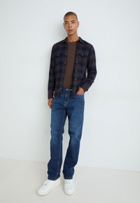 Cămașă în carouri flanelă, în nuanțe de navy și maro, purtată peste un tricou maro, asortată cu blugi albaștri și adidași albi; poziție în picioare pe un fundal simplu.